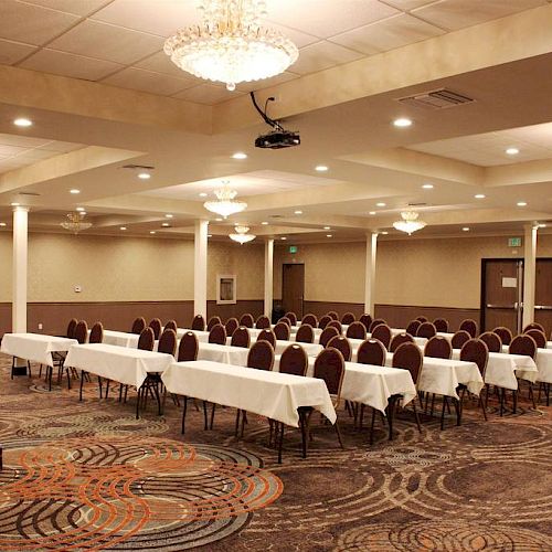 A conference room with beige walls, chandeliers, and patterned carpet. Tables with white tablecloths and rows of chairs are set up for an event.