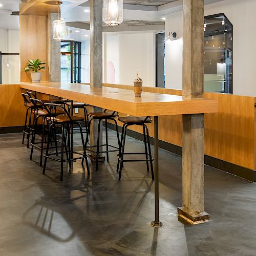 A modern café interior featuring a wooden bar-style table with black high chairs, minimalist lighting, and large windows.