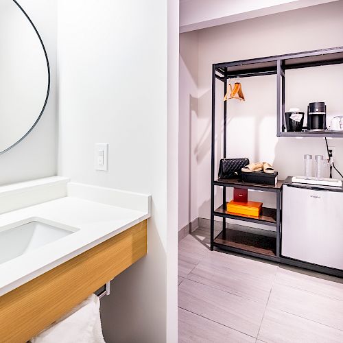 The image shows a modern bathroom sink and a kitchenette with a mini-fridge, coffee maker, and shelving unit with various items.