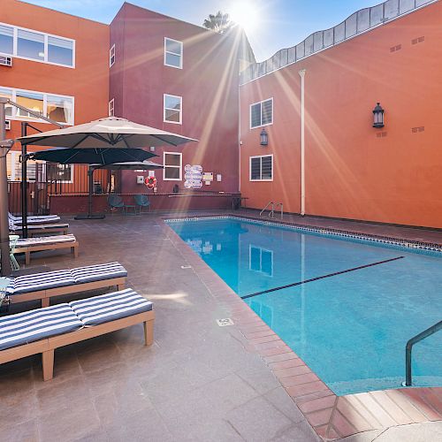 A swimming pool area with lounge chairs, umbrellas, and an orange building in the background under bright sunlight.