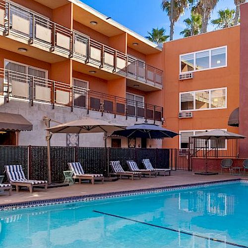 The image shows an outdoor swimming pool surrounded by lounge chairs and umbrellas, with a multi-story building and palm trees in the background.