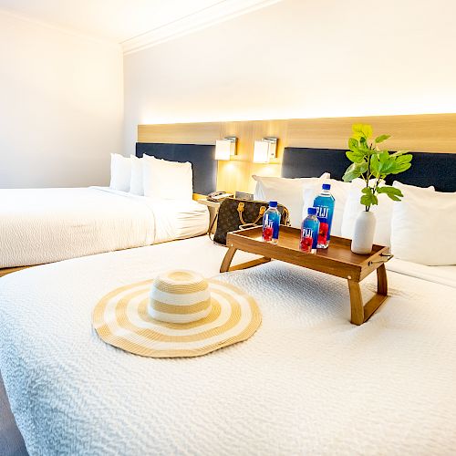 A tidy hotel room with two beds, white linens, a wooden tray with drinks, a hat, and a vase of greenery. The room has modern decor and ample lighting.