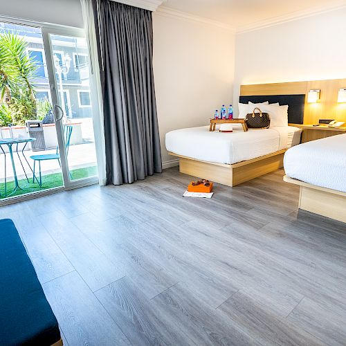 This image shows a bright hotel room with two beds, a sliding glass door leading to a patio, and modern decor including chairs and a small table.