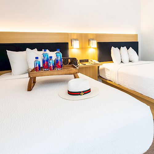 A hotel room with two beds, a wooden tray with water bottles and glasses, and a white hat on the bed, well-lit and neatly arranged.