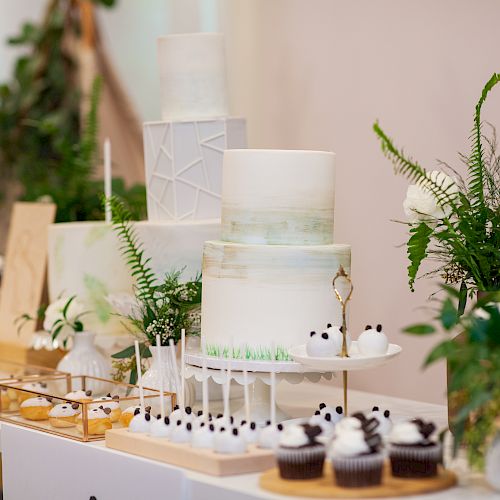 A dessert table displays elegant cakes, cupcakes, and pastries, adorned with greenery and white floral arrangements, creating a sophisticated, fresh look.