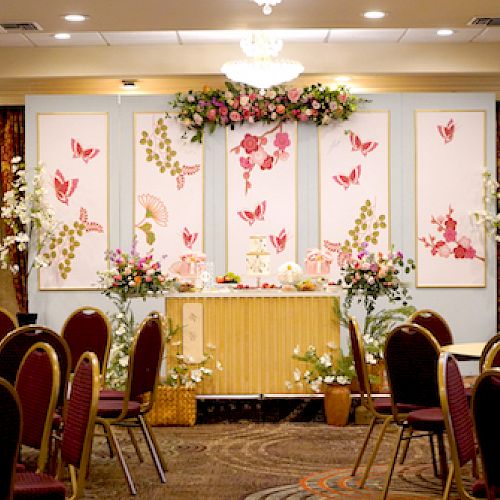 The image shows a decorated event space with round tables, chairs, and floral arrangements on a stage with butterfly-themed panels.