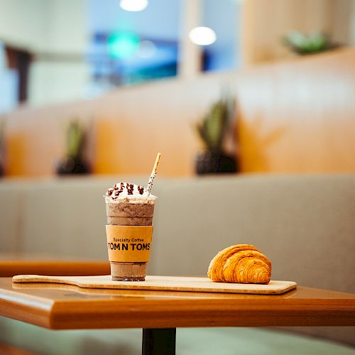 A table with a milkshake topped with whipped cream and chocolate drizzle, and a wooden board holding two croissants in a cozy indoor setting.