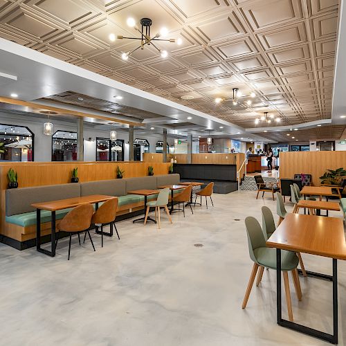 This image shows a modern, spacious dining area with wooden tables, green cushioned chairs, and booth seating, all under a decorative ceiling.