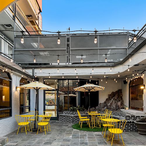 An outdoor dining area with yellow chairs, tables, and umbrellas. String lights are hung above, and there are windows on the walls.