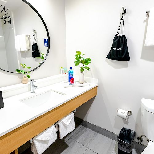 A modern bathroom with a round mirror, sink, toiletries, and towels. The decor includes a small plant, a tissue box, and a black trash bin.