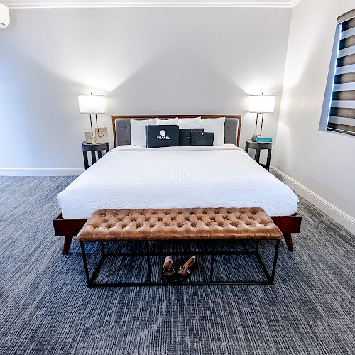 A neatly arranged bedroom with a large bed, two bedside lamps, a window with blinds, and a cushioned bench at the foot of the bed.