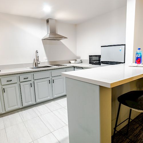 A modern kitchen with a bar counter and two barstools. Features gray cabinets, a stove, a sink, a microwave, a coffee maker, and a small refrigerator.