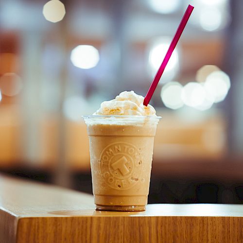 A creamy, iced coffee drink topped with whipped cream sits on a wooden surface with a red straw, in a brightly lit indoor setting.