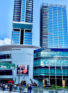 The image features two modern glass skyscrapers, a commercial building, road signs, and a crowd of people gathered near the entrance.