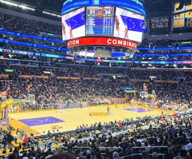 A packed arena with spectators watching a basketball game, featuring a large scoreboard hanging from the ceiling displaying the score.