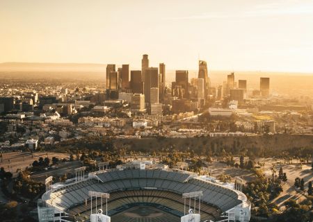 An aerial view of a stadium with a city skyline in the background during a golden sunset. The stadium is surrounded by trees and parking lots.