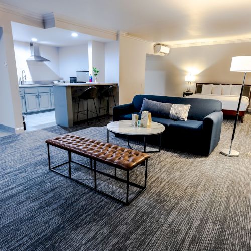 A modern living space features a blue sofa, round coffee table, bench, floor lamp, and kitchen area with bar stools and overhead cabinets.