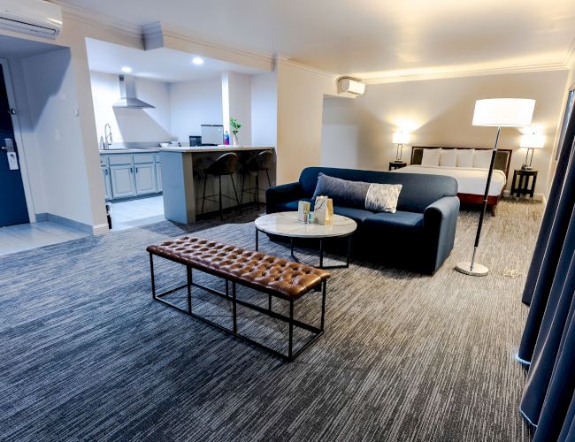 A modern living space features a blue sofa, round coffee table, bench, floor lamp, and kitchen area with bar stools and overhead cabinets.