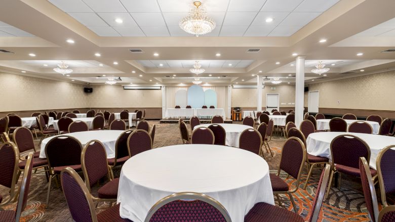 The image shows a large banquet hall with round tables covered with white cloths and surrounded by chairs, and chandeliers hanging from the ceiling.