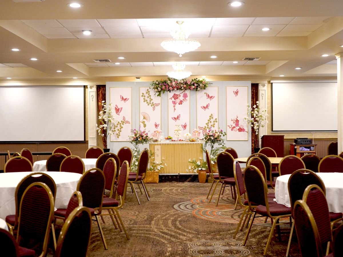 A decorated event hall with round tables, red chairs, floral arrangements, two projection screens, and a chandelier on the ceiling.