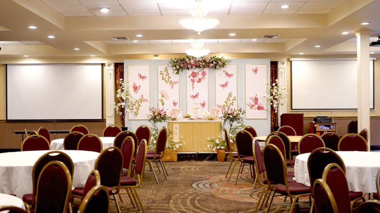 A decorated event hall with round tables, red chairs, floral arrangements, two projection screens, and a chandelier on the ceiling.