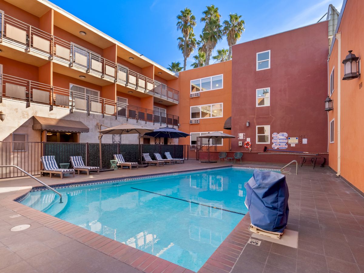 An outdoor swimming pool with lounge chairs and umbrellas, surrounded by a multi-story hotel or apartment building on a clear, sunny day.