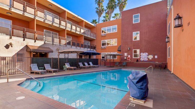 An outdoor swimming pool with lounge chairs and umbrellas, surrounded by a multi-story hotel or apartment building on a clear, sunny day.