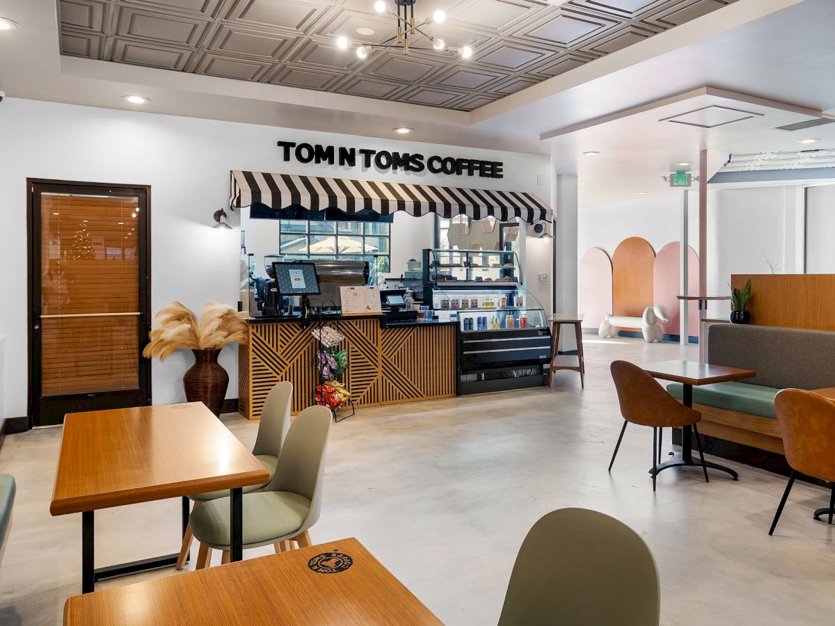 A modern cafe with "Tom N Toms Coffee" signage, wooden tables, green and brown chairs, decorative plants, and a polished, stylish interior finish.