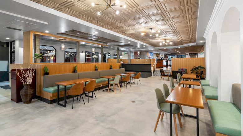 A modern, spacious dining area with wooden tables, green upholstered seating, and minimalist décor is depicted in this image.