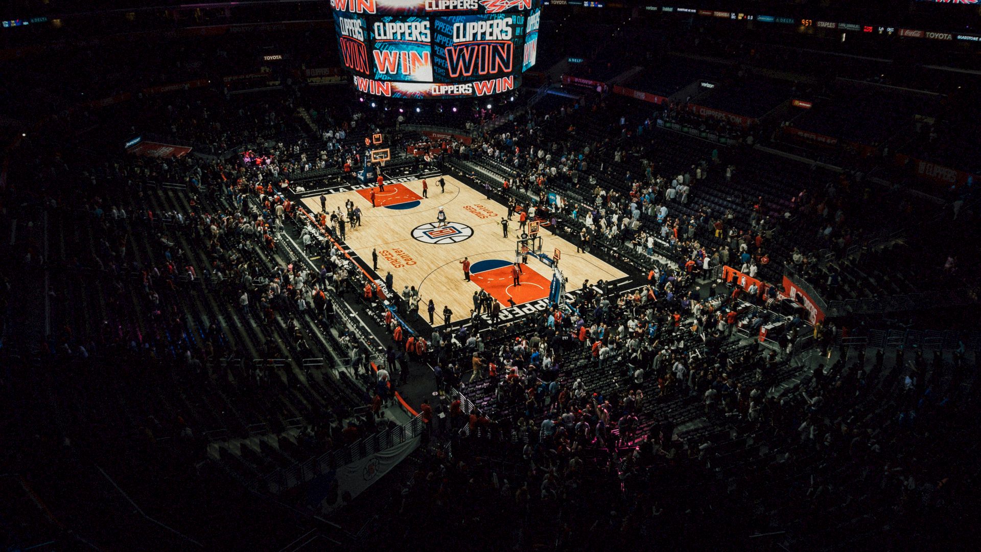 A basketball arena shows "Clippers Win" on the screen. Fans are present, and the court is illuminated with team logos.