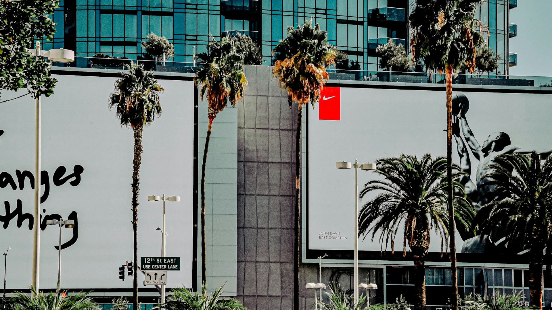 The image shows two large billboards on a building with palm trees in front. One is partially visible, and the other features a basketball player.