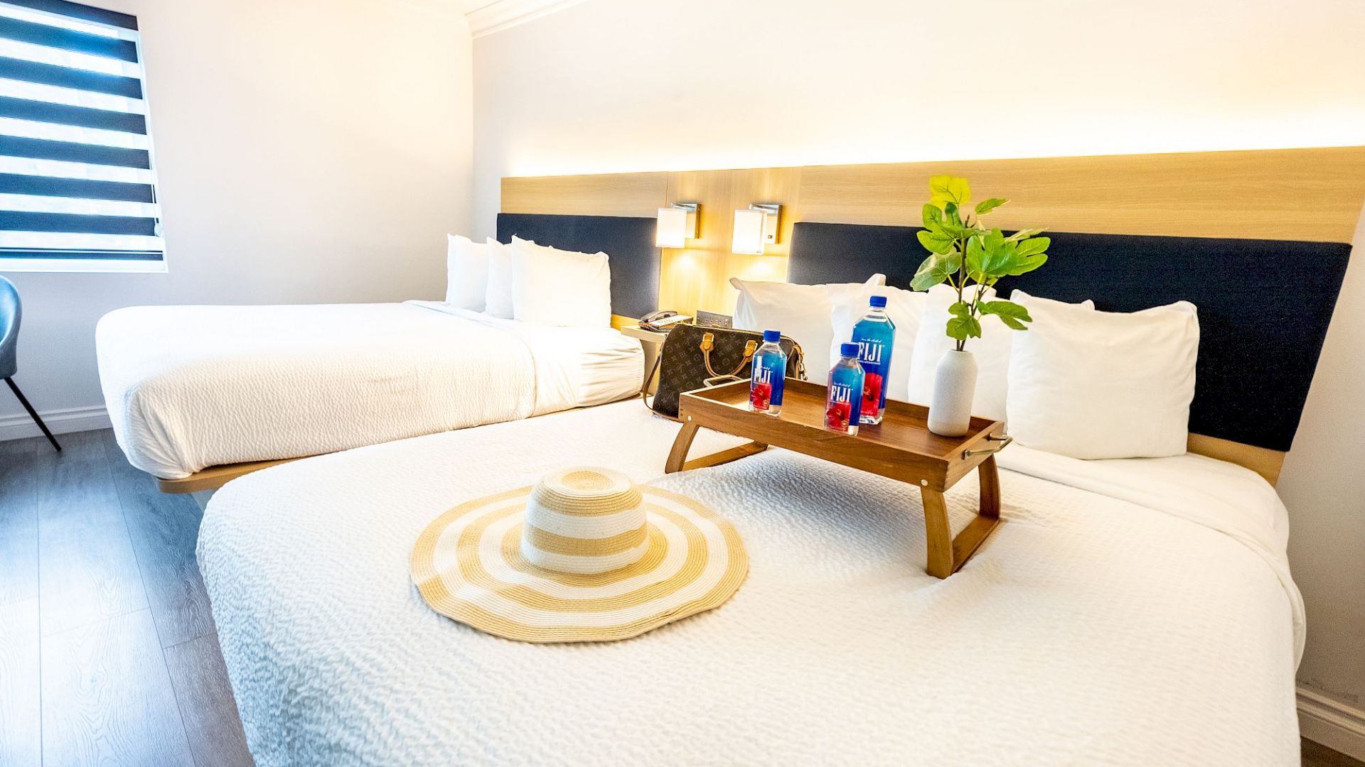 The image shows a bright hotel room with two neatly made beds, a hat, a wooden tray holding drinks, and decor items.