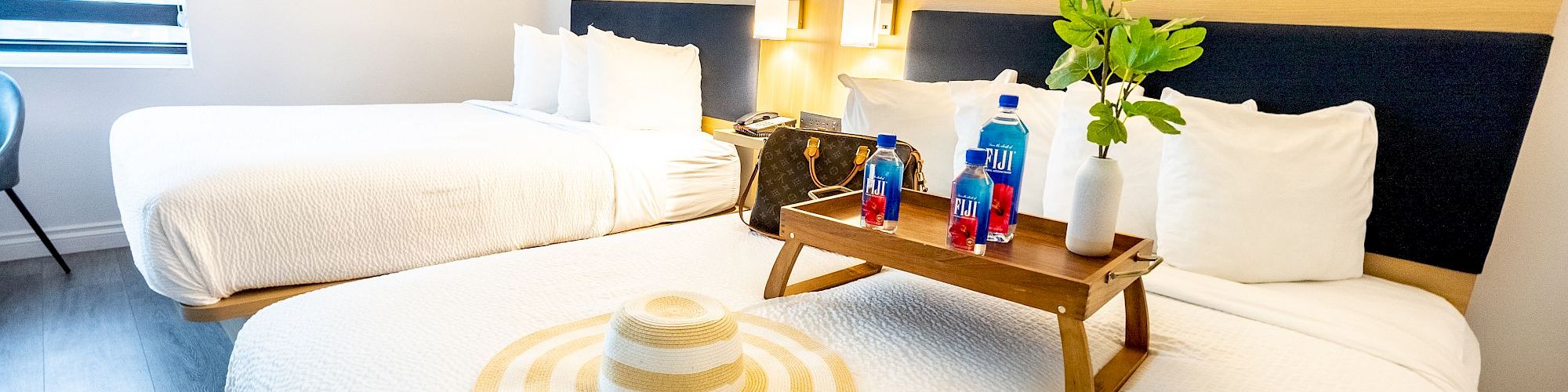 The image shows a bright hotel room with two neatly made beds, a hat, a wooden tray holding drinks, and decor items.