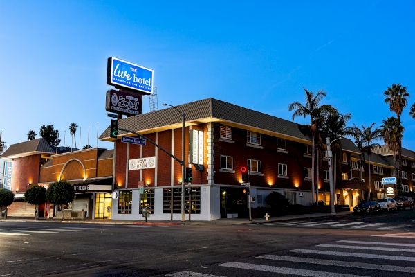 A brick building with a "Live Hotel" sign is on a street corner at dusk, with palm trees and street lights illuminating the area.