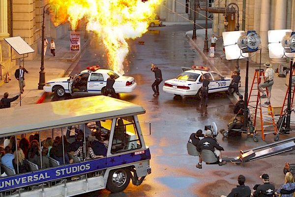 The image shows a movie set with an explosion, police cars, film crew, lighting equipment, and a Universal Studios tour bus on the street.