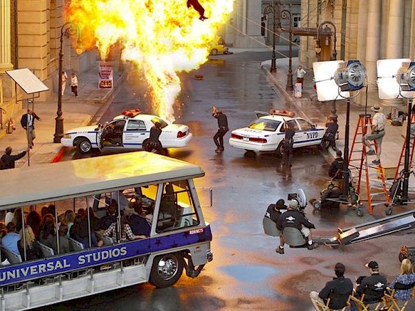 The image shows a movie set with an explosion, police cars, film crew, lighting equipment, and a Universal Studios tour bus on the street.
