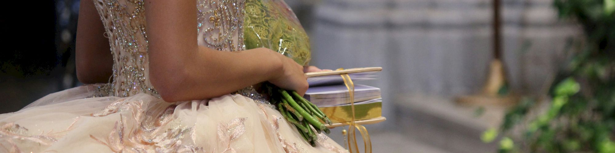 A person in an elaborate, beaded gown holds a bouquet and a wrapped gift, standing in what appears to be a formal setting.