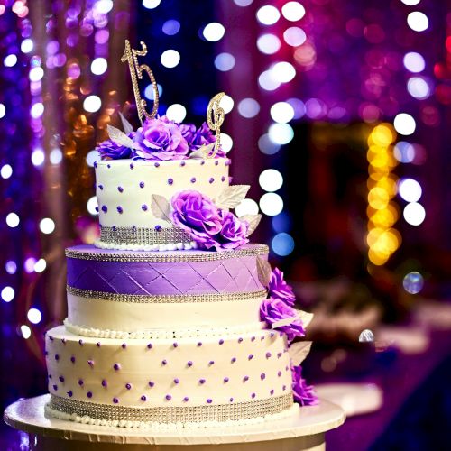 A three-tiered wedding cake adorned with purple flowers and decorations, set against a background of colorful, out-of-focus lights.