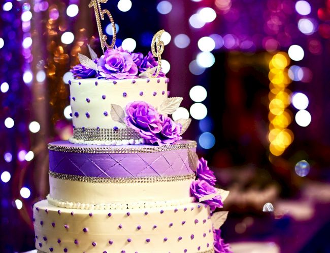 A three-tiered wedding cake adorned with purple flowers and decorations, set against a background of colorful, out-of-focus lights.