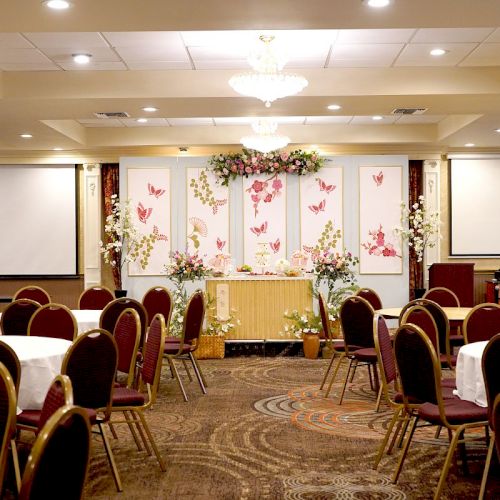 The image shows a banquet hall with round tables and chairs, a decorated stage, and two projection screens. The hall is prepared for an event.