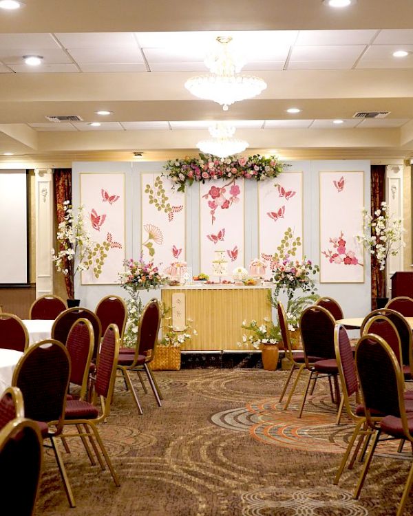 The image shows a banquet hall with round tables and chairs, a decorated stage, and two projection screens. The hall is prepared for an event.