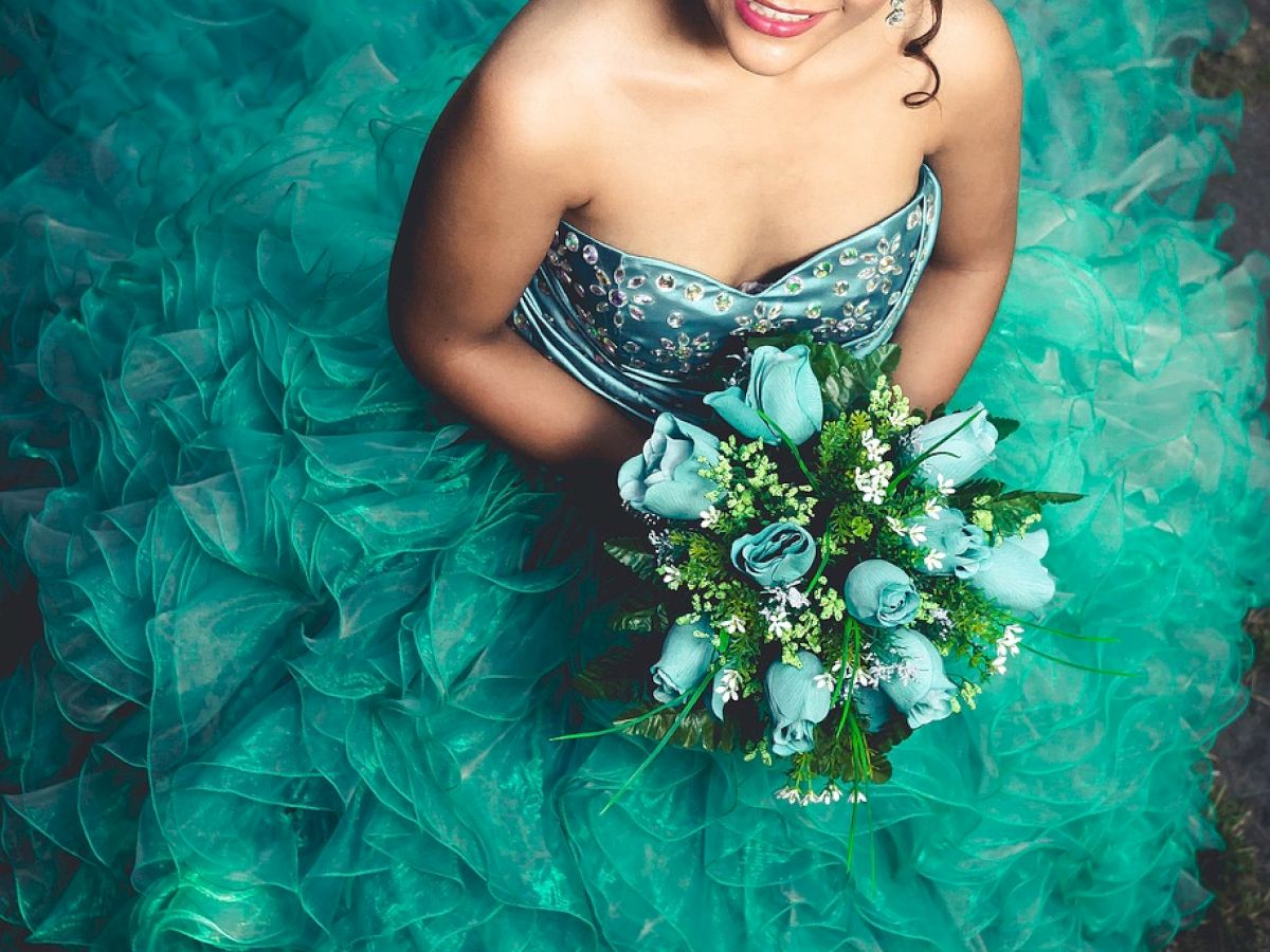 A woman in a strapless teal dress, holding a bouquet of blue and white flowers, smiles while looking up at the camera.