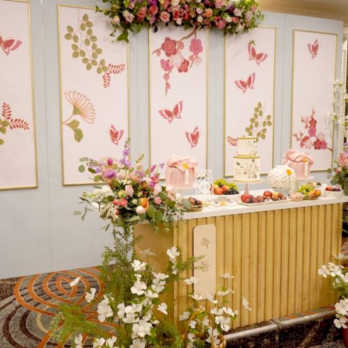 The image shows a beautifully decorated table with flowers, a cake, and fruits, set against a backdrop with butterfly and floral designs.