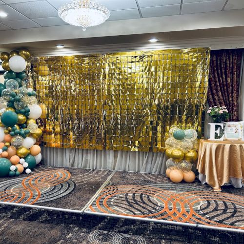 The image shows a decorated room with a gold fringe backdrop, balloon arrangements, and a table with a cloth, flowers, and a letter "E" standing on it.