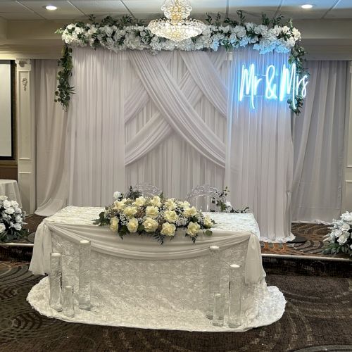 The image shows a wedding reception setup with a decorated table, white drapes, flowers, and a 