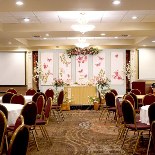 The image shows a decorated banquet hall with round tables and chairs, floral decorations, and projection screens, likely set up for an event.