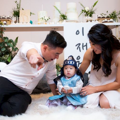 A family is sitting together on a cozy rug, with the child dressed in traditional attire, and decorative items and plants in the background.