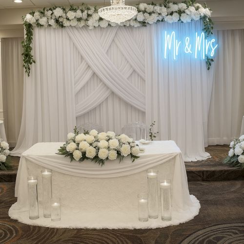 A decorated wedding table with white drapes, floral arrangements, candle holders, and a neon "Mr & Mrs" sign completes the elegant setup.