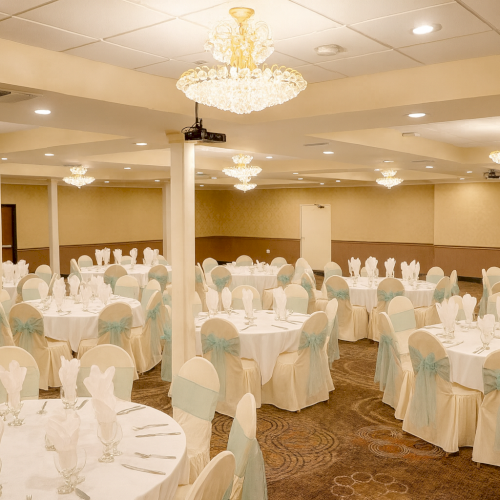 The image shows an elegant banquet hall with round tables, white napkins, and elegant chandeliers, prepared for a formal event or reception.
