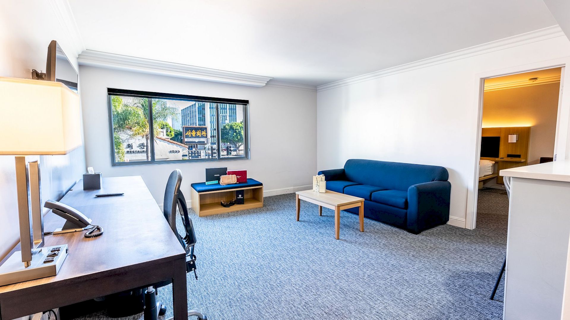 A bright living room with a blue couch, desk, chair, table, and window view. Adjoining room visible in the background.